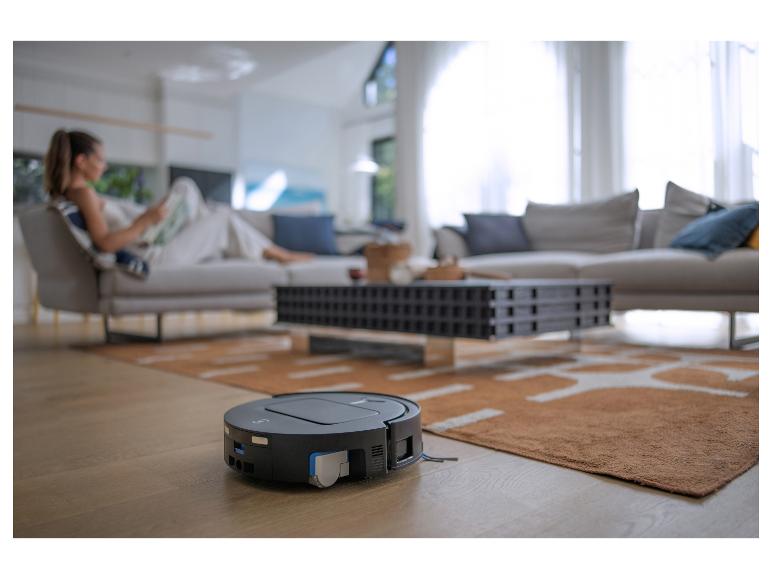 Saugroboter auf Holzboden im Wohnzimmer mit lesender Frau im Hintergrund.
