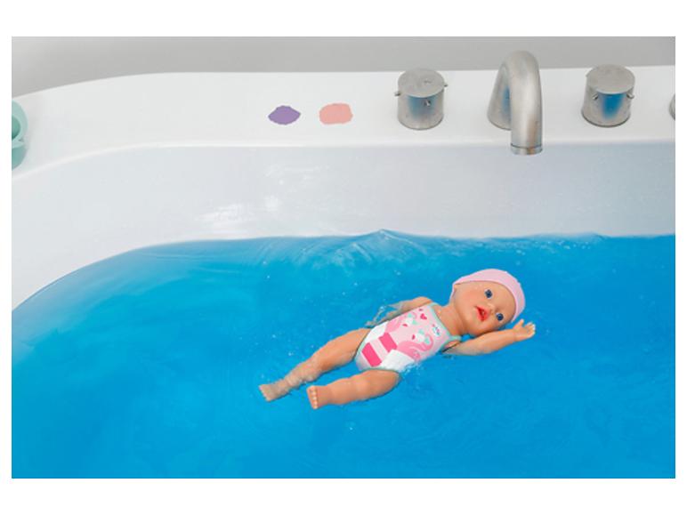 Babypuppe schwimmt in blauem Wasser in einer Badewanne, trägt einen rosa Badeanzug.