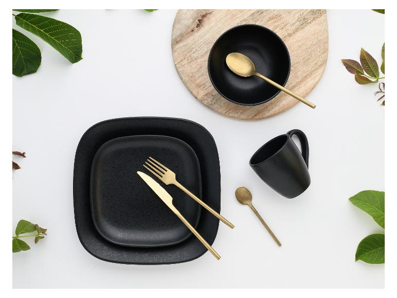 Schwarzes Geschirrset mit goldenem Besteck, Tasse und Schale auf einem Holzbrett, mit grünen Blättern.