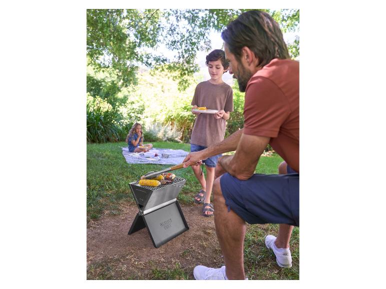 Vater und Sohn grillen auf einem tragbaren Grill, Mädchen sitzt auf einer Decke im Hintergrund.
