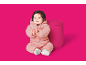 Glückliches Baby in einem rosa gepolsterten Schneeanzug, klatscht in die Hände neben einem rosa Pouf.