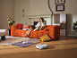 Frau auf orangefarbenem Sofa mit Laptop, Saugroboter auf Holzfußboden.