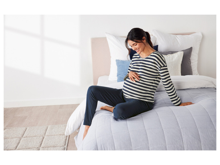 Eine schwangere Frau in einem gestreiften Pyjama und blauen Hosen sitzt auf einem Bett.