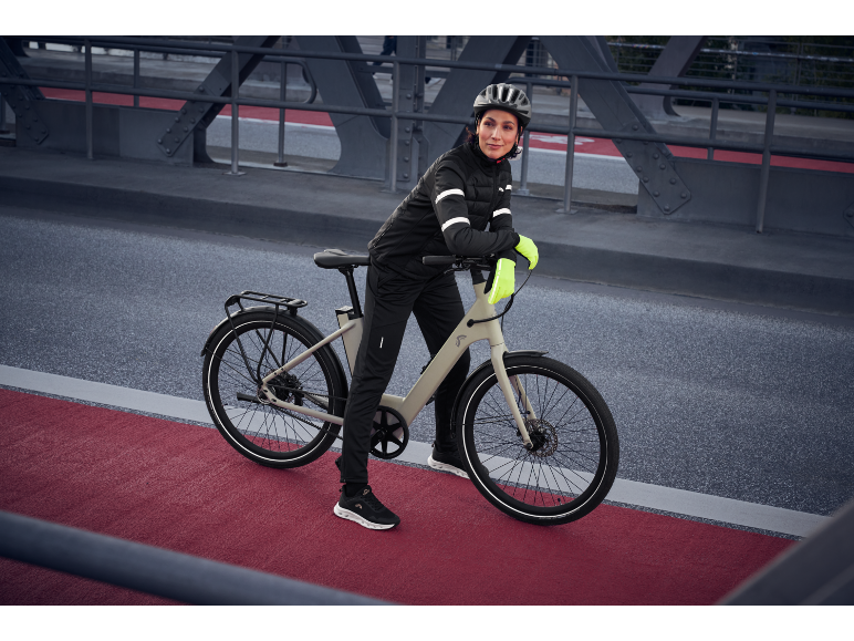 Frau in schwarzer Radsportbekleidung und gelben Handschuhen mit E-Bike auf einer Brücke.