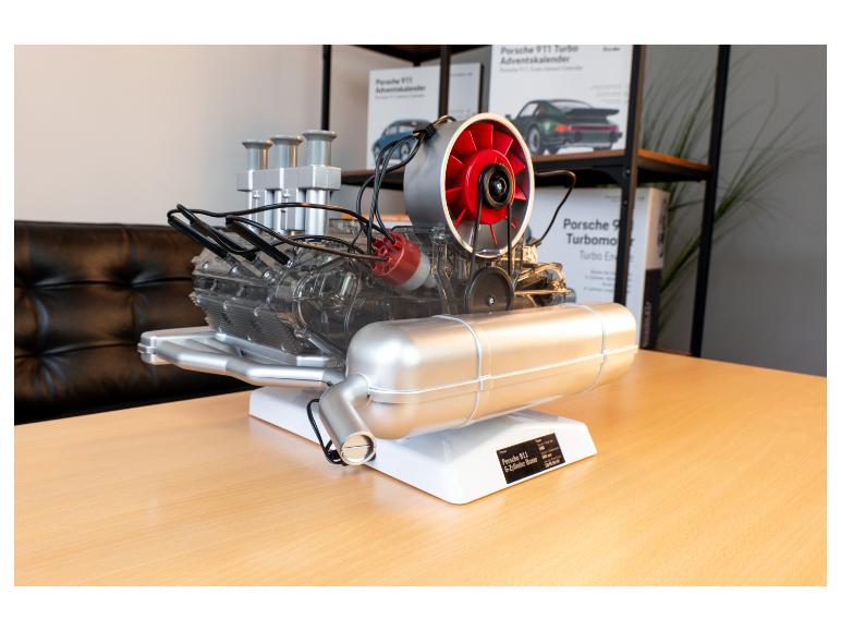 Porsche 911 Motormodell auf einem Tisch, mit Porsche-Boxen im Hintergrund.