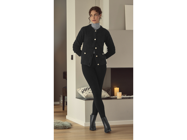 Frau in schwarzer Strickjacke, Rollkragenpullover und Leggings, posiert in einem gemütlichen Raum.