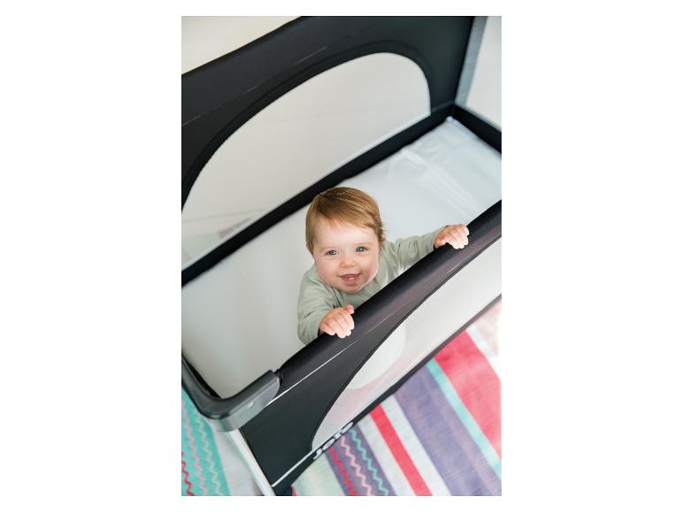 Lächelndes Baby in einem schwarz-weißen Reisebett mit buntem Teppich.