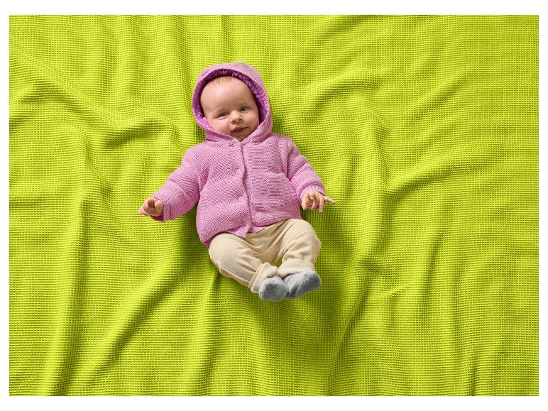 Baby in rosa Kapuzenpullover und beiger Hose auf einer lindgrünen Waffeldecke.