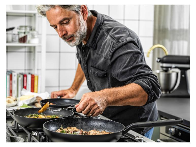 Mann brät Fleisch und Kartoffeln in schwarzen Pfannen auf einem Gasherd