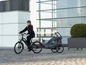 Eine Frau fährt mit einem E-Bike, an dem ein Qeridoo-Kinderanhänger befestigt ist.