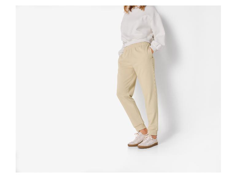 Frau trägt beiges Sweatshirt und beige Jogginghose auf weißem Hintergrund.