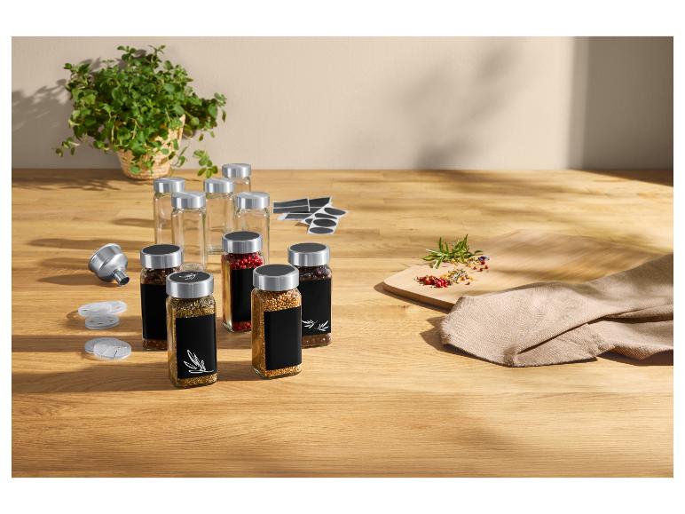 Gewürze in Glasgefäßen mit schwarzen Etiketten, Trichter und frische Kräuter auf Holztisch.
