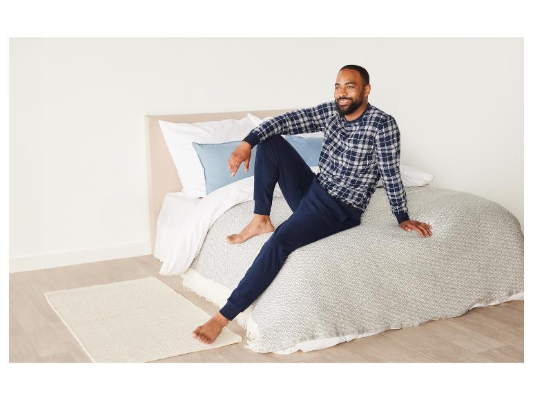 Mann in kariertem Pyjama sitzt auf einem Bett mit einem weißen Überwurf.