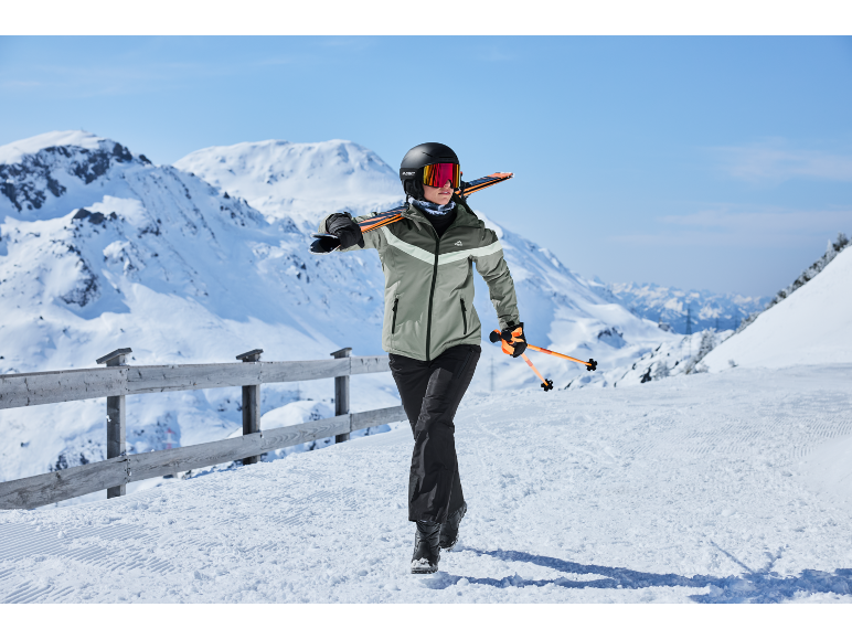 Frau in Skijacke, Hose, Helm und Brille trägt Skier und Stöcke vor verschneiter Bergkulisse.
