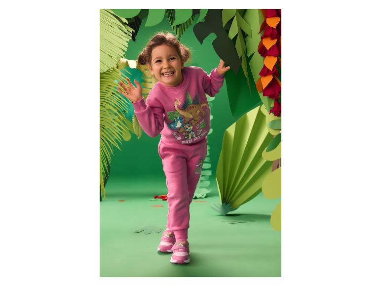 Mädchen in pinkfarbenem Trainingsanzug mit Dinosaurier-Print und Turnschuhen, lächelnd in einer Papierdschungelkulisse.