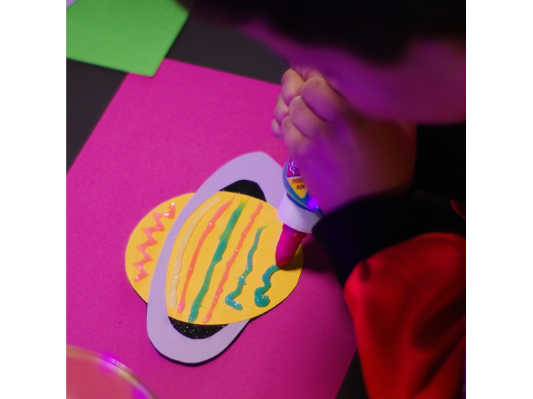 Kind verziert ein Papierbastelprojekt mit farbigem Glitzerkleber auf rosa Hintergrund.