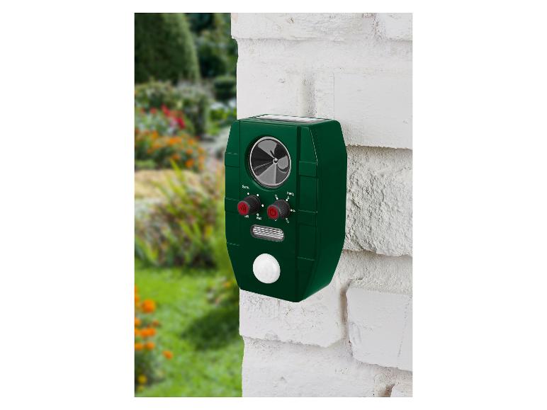 Grüner Tiervertreiber mit Solarpanel an einer weißen Ziegelwand im Garten.