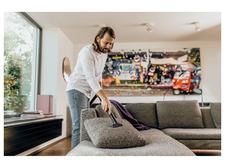 Mann saugt Sofa mit einem Handstaubsauger im Wohnzimmer.