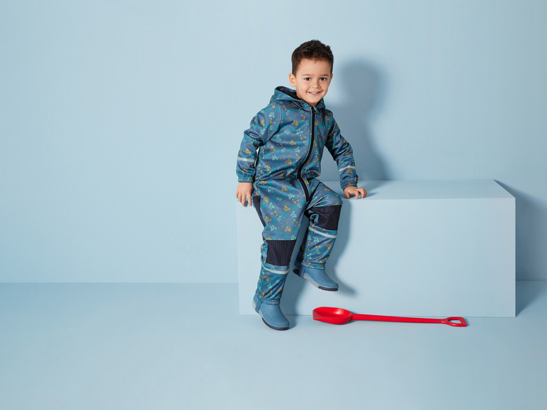 Ein Kind in einem blauen Overall mit Fahrrad-Print und blauen Stiefeln sitzt auf einer Plattform mit einer roten Schaufel.