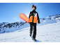 Junge Person in Skijacke und Hose mit Snowboard am verschneiten Berghang.