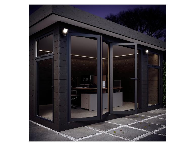 Modernes Gartenbüro mit großen Glastüren, Innenbeleuchtung und Außenstrahlern bei Nacht.
