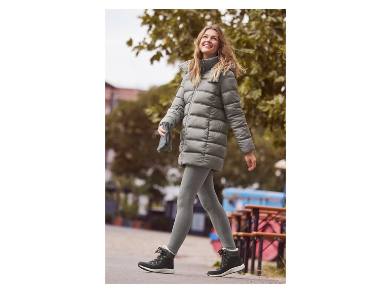 Eine Frau in einer grauen Winterjacke, Leggings und schwarzen Stiefeln geht auf der Straße.