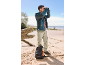Mann blickt mit Fernglas am Strand, trägt Outdoor-Jacke und -Hose, Rucksack daneben.