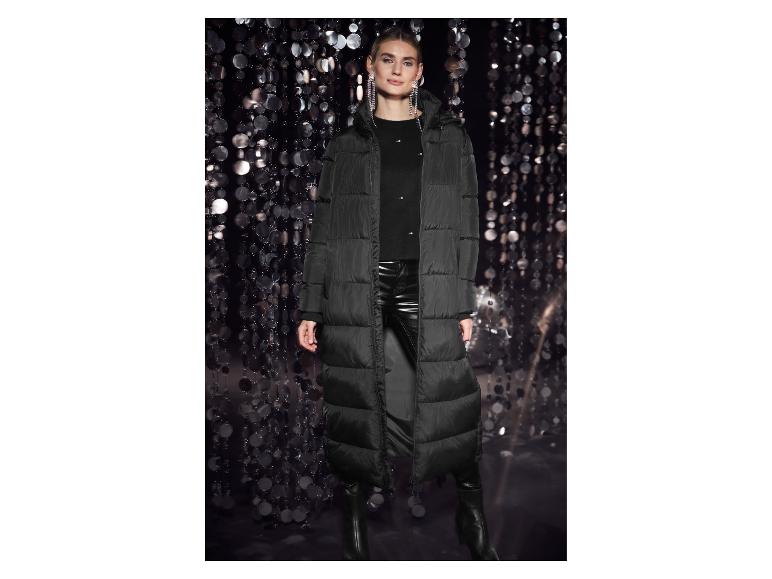 Frau in schwarzem langen Daunenmantel, schwarzen Lederhosen und Stiefeln vor glänzendem Hintergrund.