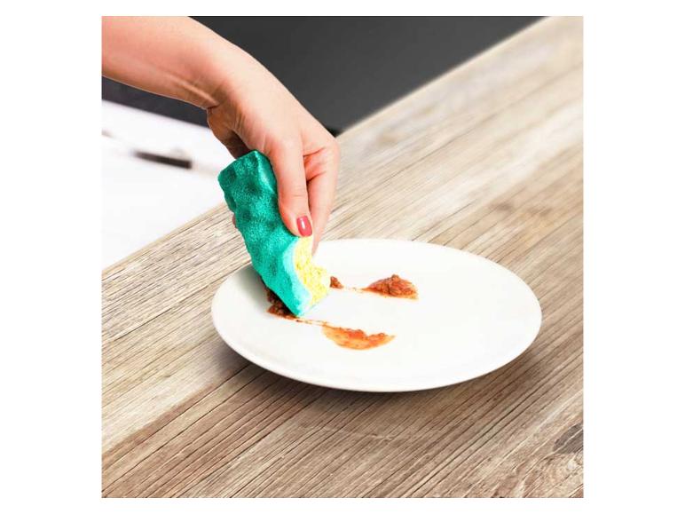 Hand reinigt Teller mit Spülschwamm auf Holztisch.