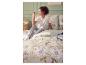 Frau im Pyjama sitzt auf einem Bett mit floraler Bettwäsche, daneben ein Nachttisch mit Büchern und Tasse.