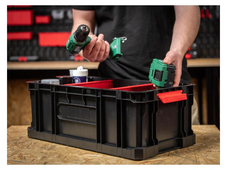 Ein Mann legt einen grünen Bohrer und Akku in eine schwarze Aufbewahrungsbox mit roten Fächern.