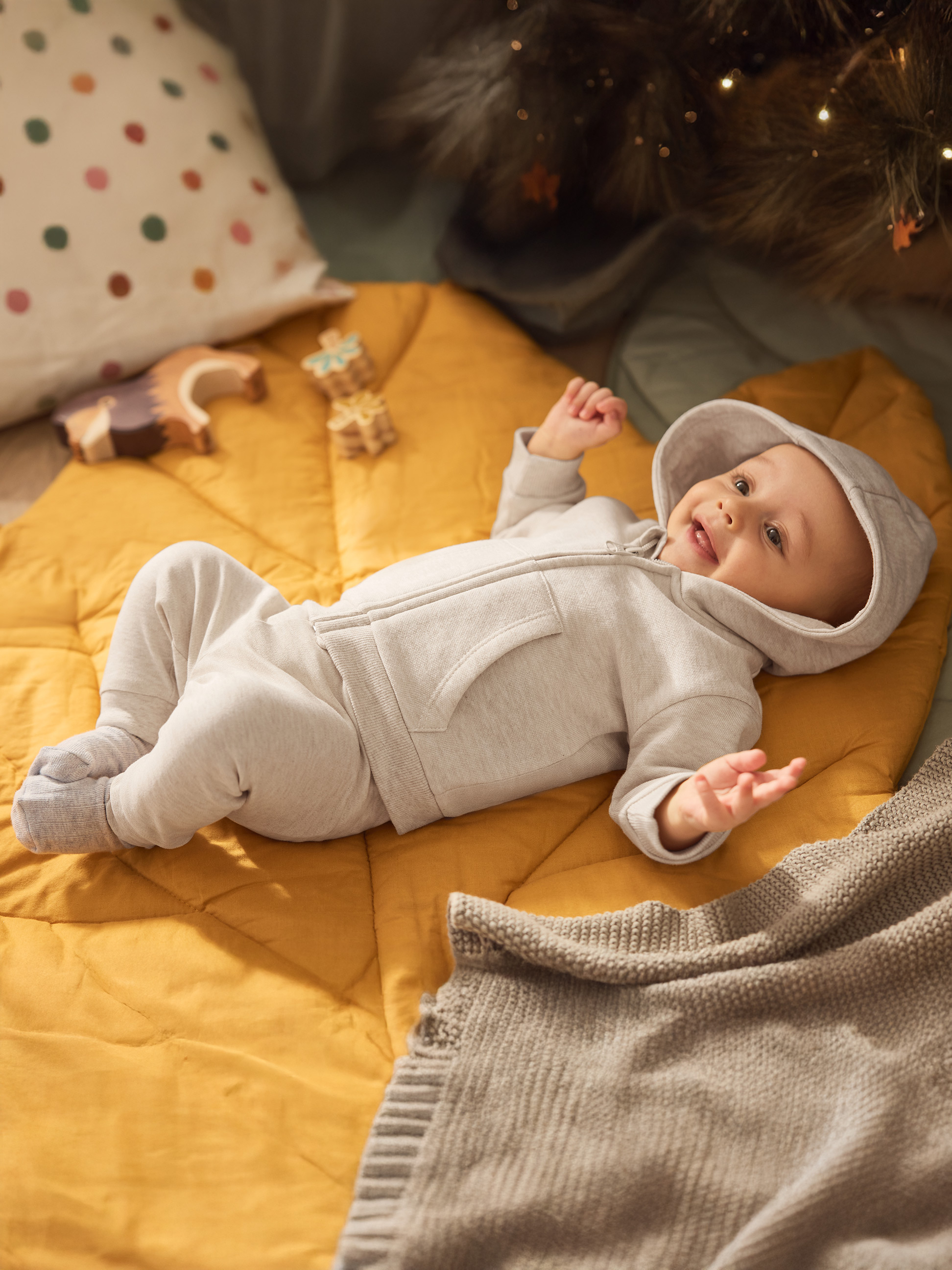 Glückliches Baby im grauen Strampler auf gelber Matratze mit Spielzeug und Weihnachtslichtern.