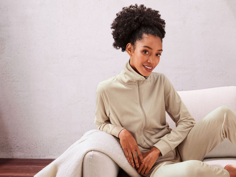 Eine Frau in einem beigen Trainingsanzug sitzt auf einem Sofa.