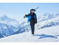 Mann in Skijacke, Hose, Helm und Brille trägt Skier und Stöcke auf verschneitem Berg.