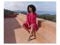 Frau in fuchsiafarbenem Kleid und bequemen Schuhen sitzt auf einer Terrasse mit Meerblick.