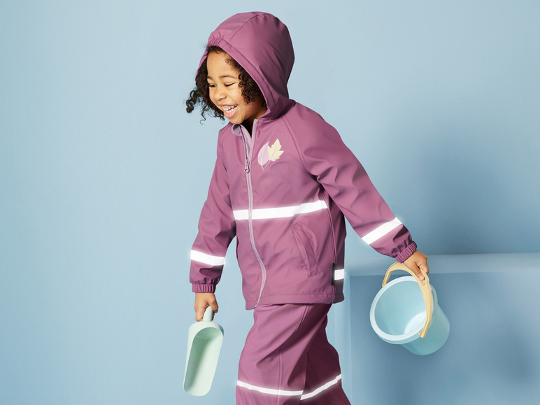 Ein Kind trägt eine lila Regenjacke und -hose mit reflektierenden Streifen und hält eine Schaufel und einen Eimer.