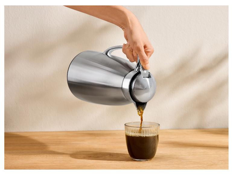 Hand gießt Kaffee aus einer silbernen Thermoskanne in ein Glas auf einem Holztisch.