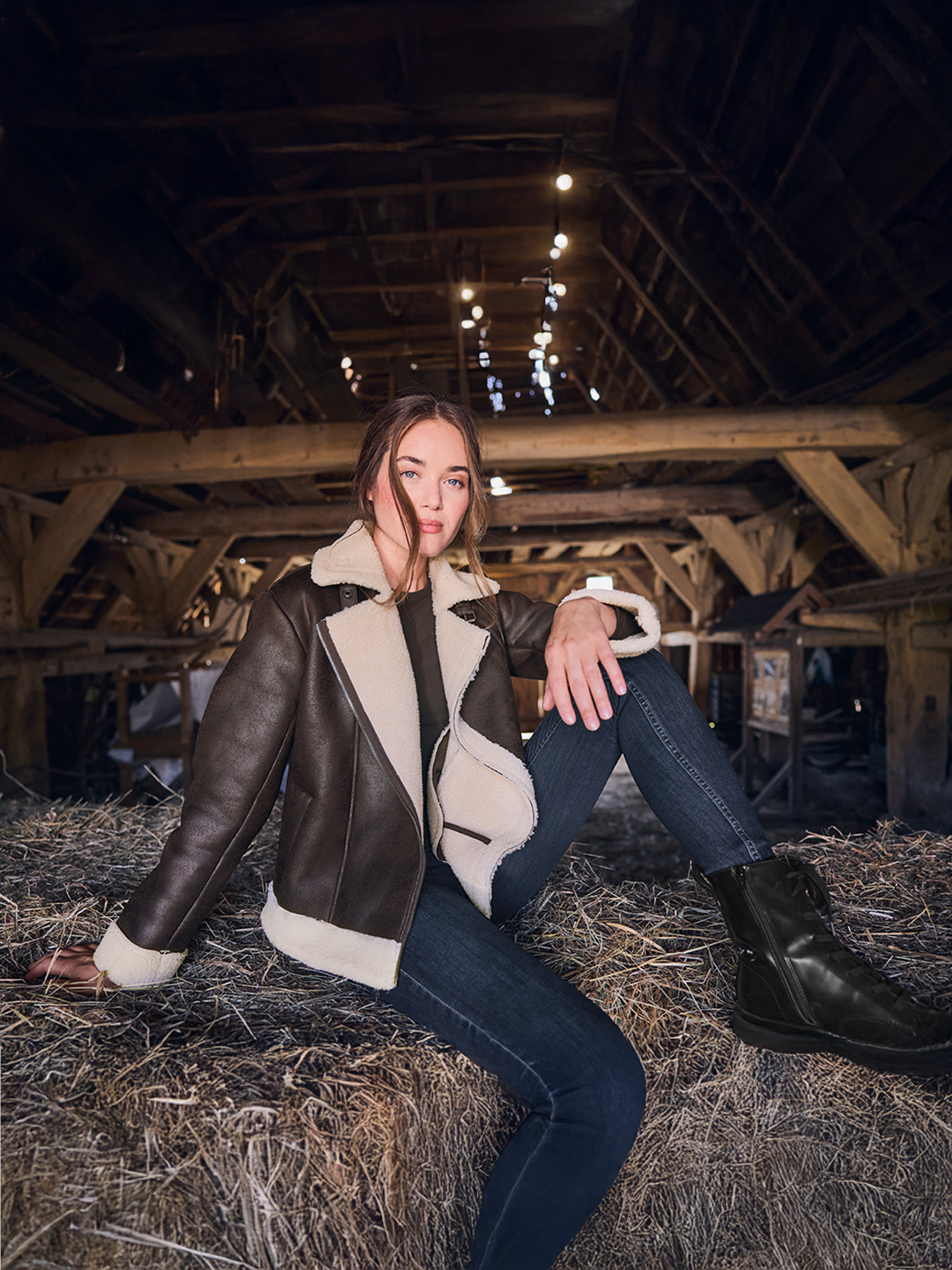 Frau in brauner Shearling-Jacke und Jeans, auf Heuballen in einer Scheune sitzend.