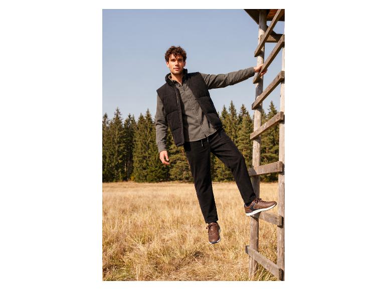 Mann in Jeans, Weste und braunen Sneakern auf einer Holzleiter in der Natur.