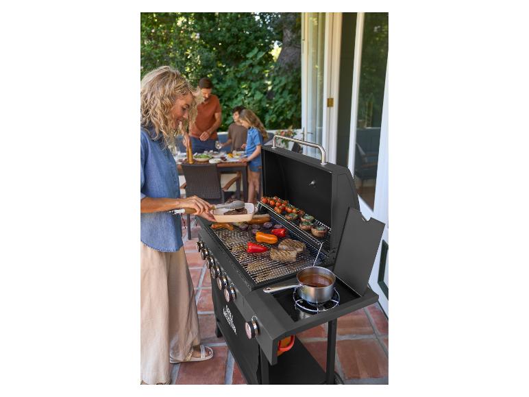 Frau grillt Fleisch und Gemüse auf einem Gasgrill, im Hintergrund Familie am Tisch auf der Terrasse.