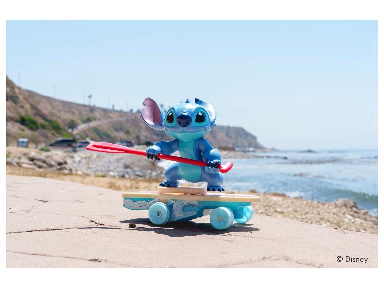 Stitch-Figur auf Skateboard mit Paddel, auf einem Weg am Strand.