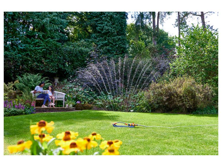 Ein Rasensprenger bewässert einen grünen Rasen, während zwei Personen auf einer Gartenbank sitzen.