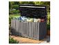 Graue Outdoor-Aufbewahrungsbox mit bunten Kissen auf einer Holzterrasse in einem blühenden Garten.
