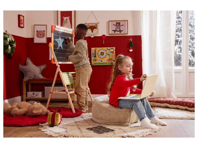 Zwei Kinder spielen in einem weihnachtlich dekorierten Zimmer: ein Junge zeichnet an einer Tafel, ein Mädchen benutzt einen Laptop.