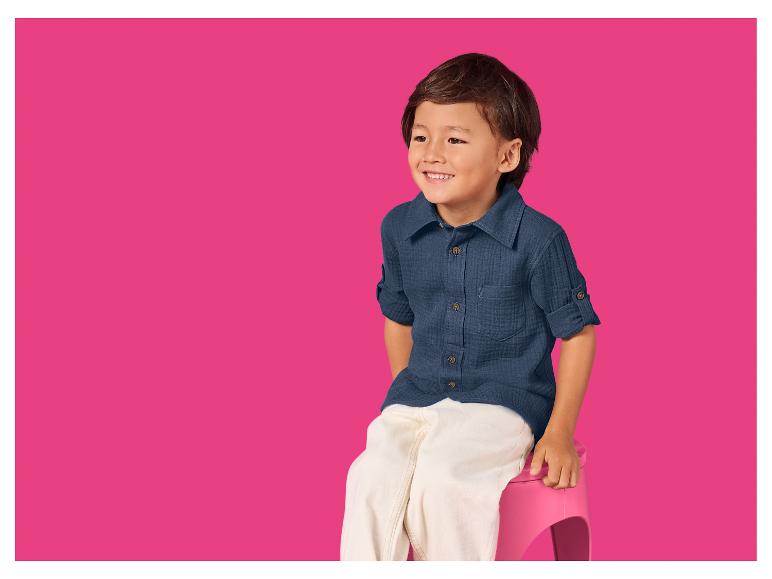 Lächelnder Junge in blauem Hemd und weißer Hose sitzt auf einem rosa Hocker.