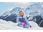 Mädchen in Winteranzug und Handschuhen, lachend im Schnee mit Bergen im Hintergrund.