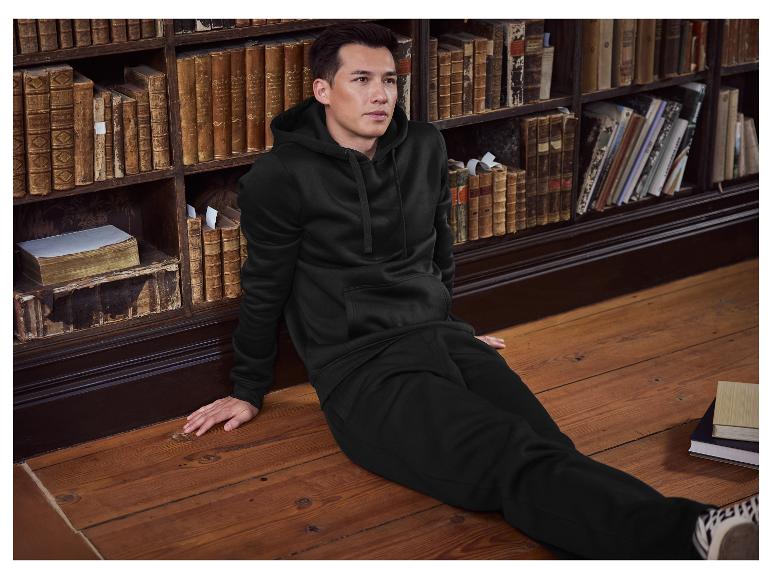 Mann in schwarzem Hoodie und Jogginghose sitzt auf dem Boden neben einem Bücherregal.