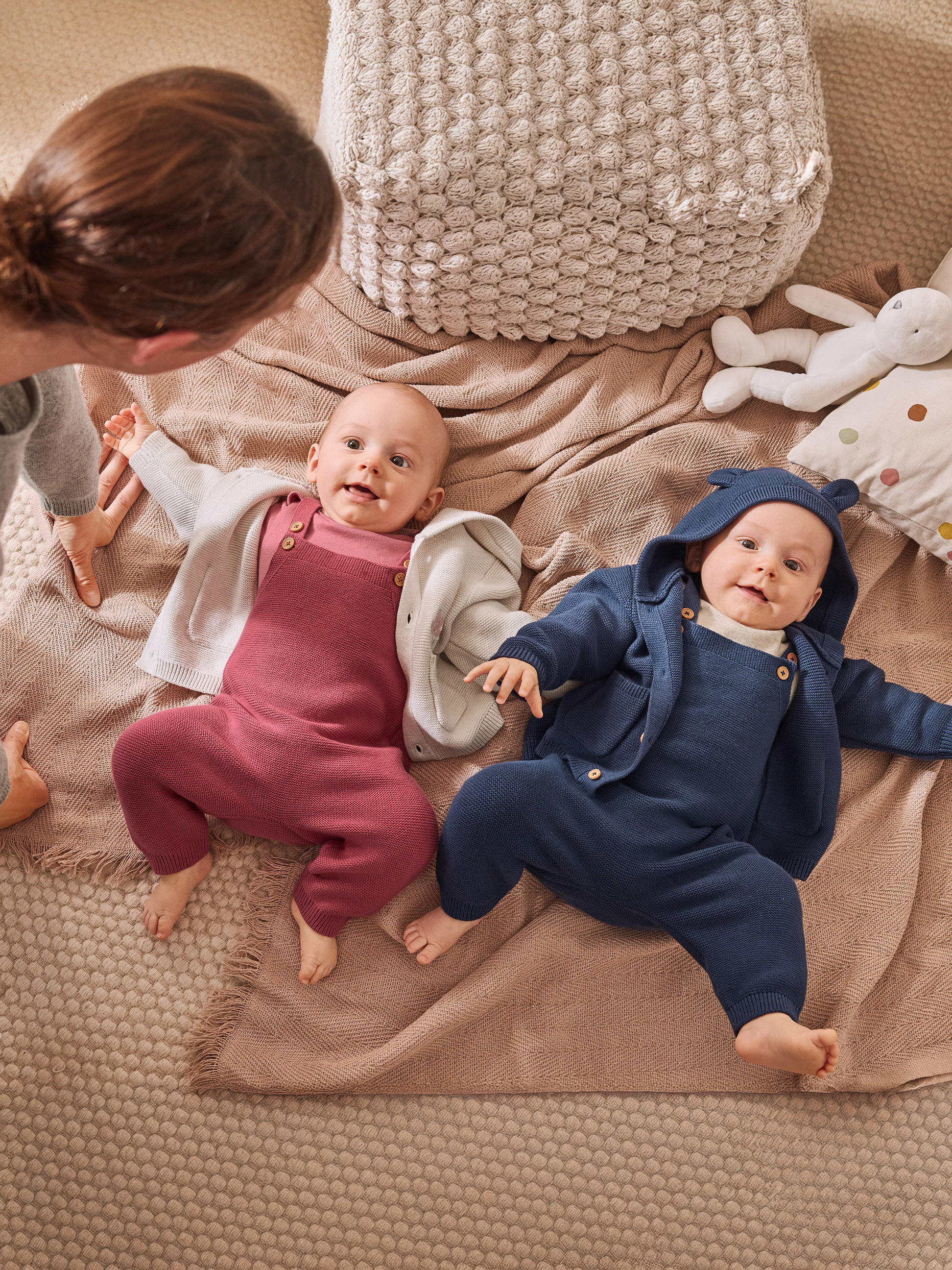 Zwei lächelnde Babys in gestrickten Overalls, einer rosa und einer blau, auf einer Decke.