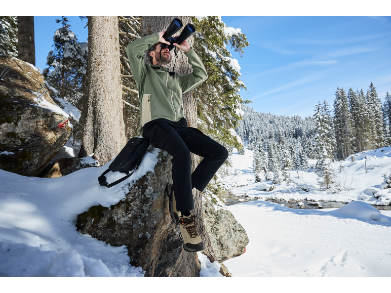 Mann in Winterjacke und Hose sitzt auf einem Felsen und schaut mit Fernglas in verschneitem Wald.
