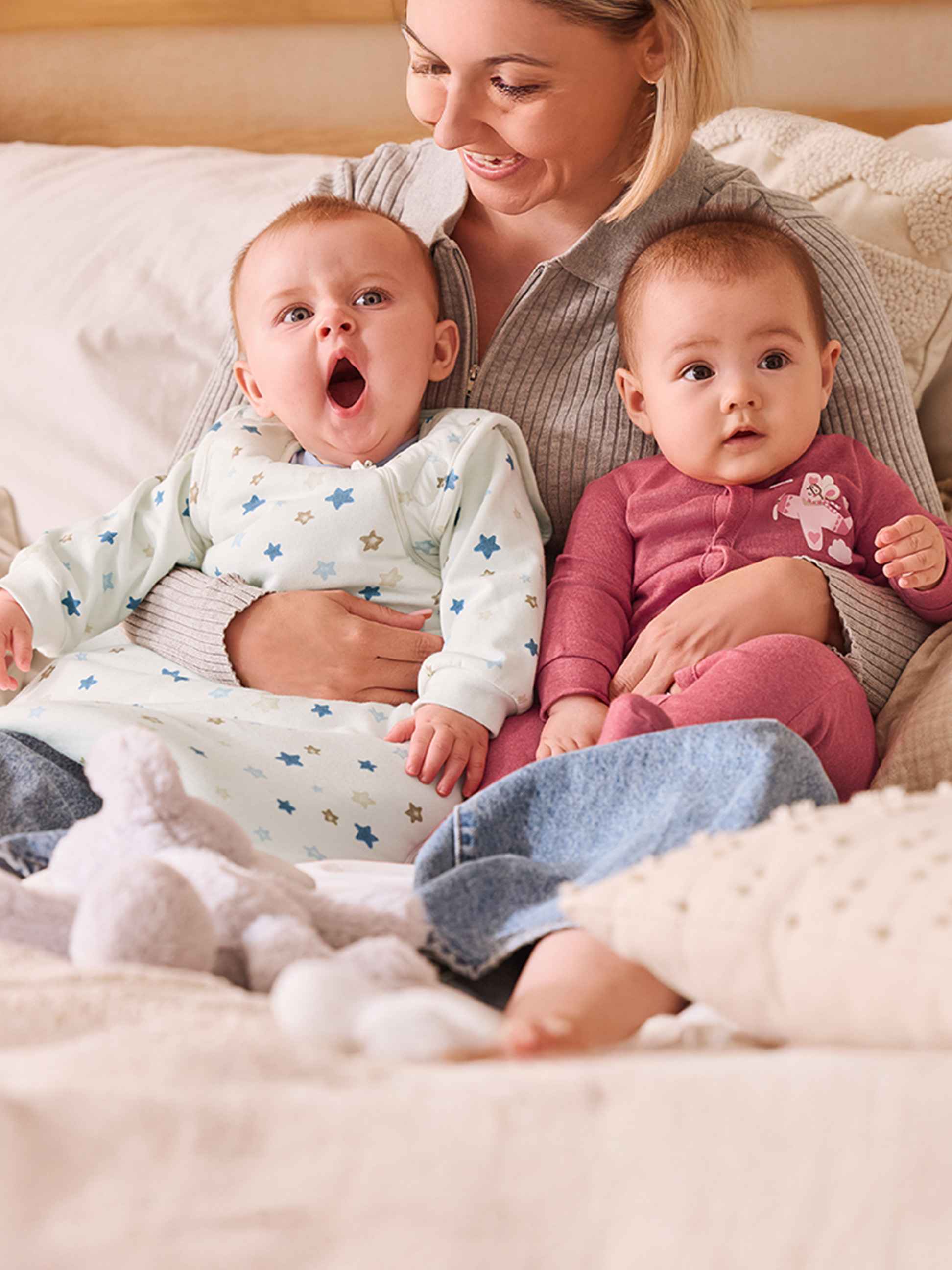 Eine lächelnde Mutter hält zwei Babys, eines gähnt, das andere schaut neugierig.